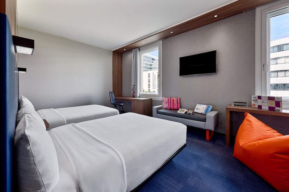 A hotel room with two beds, a desk, a TV, and a window with a city view.