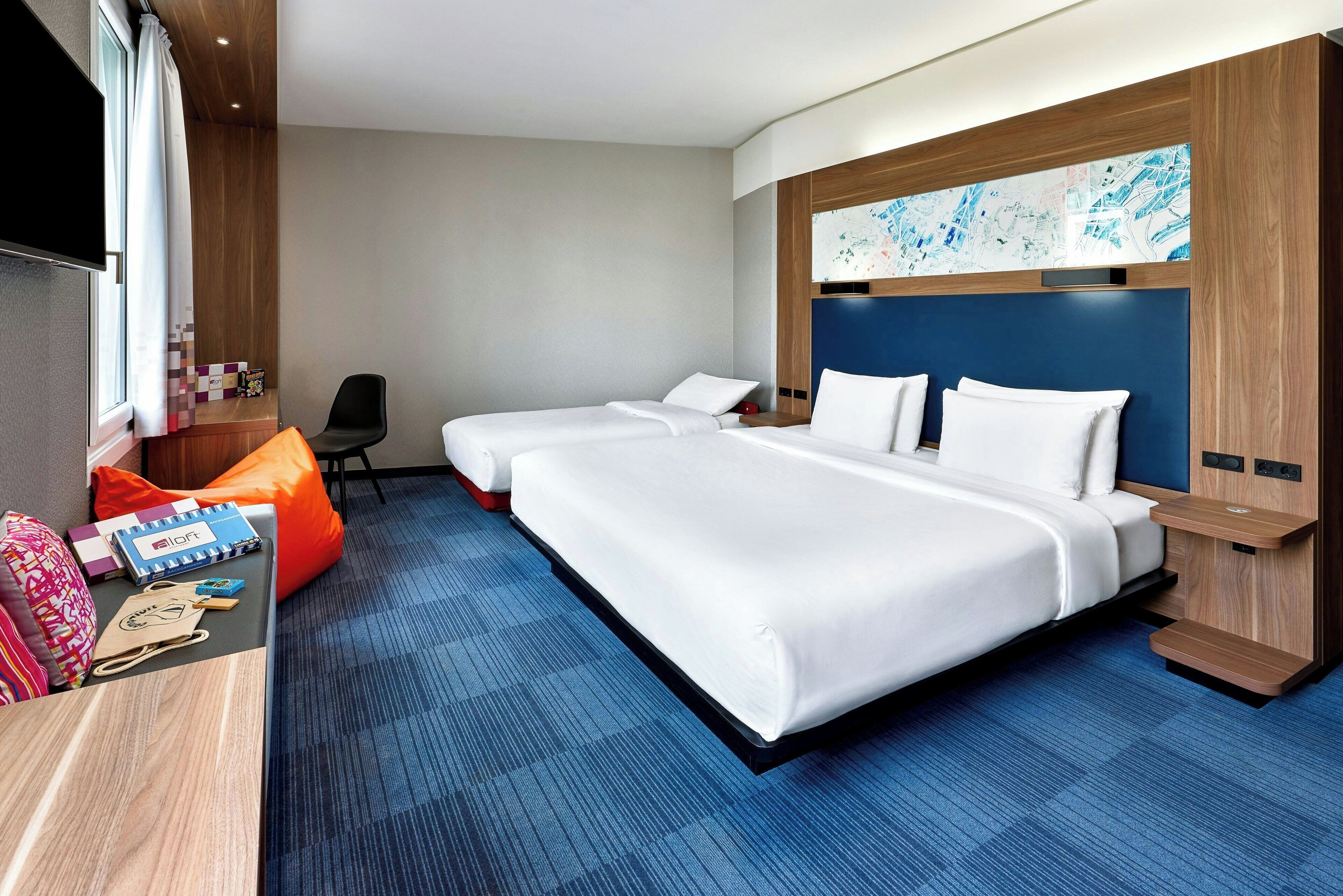 A hotel room with two beds, a desk with a chair, and a large window with curtains.