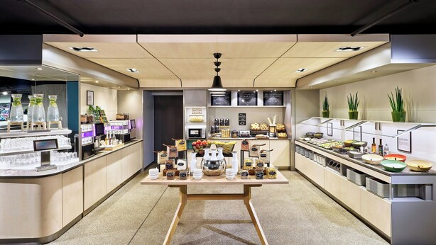 A modern café with a central table displaying various food items and beverages, surrounded by shelves with snacks and condiments.