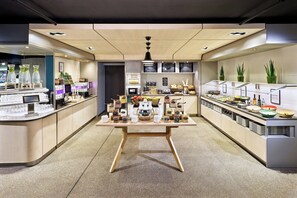 A modern café with a central table displaying various food items and beverages, surrounded by shelves with snacks and condiments.