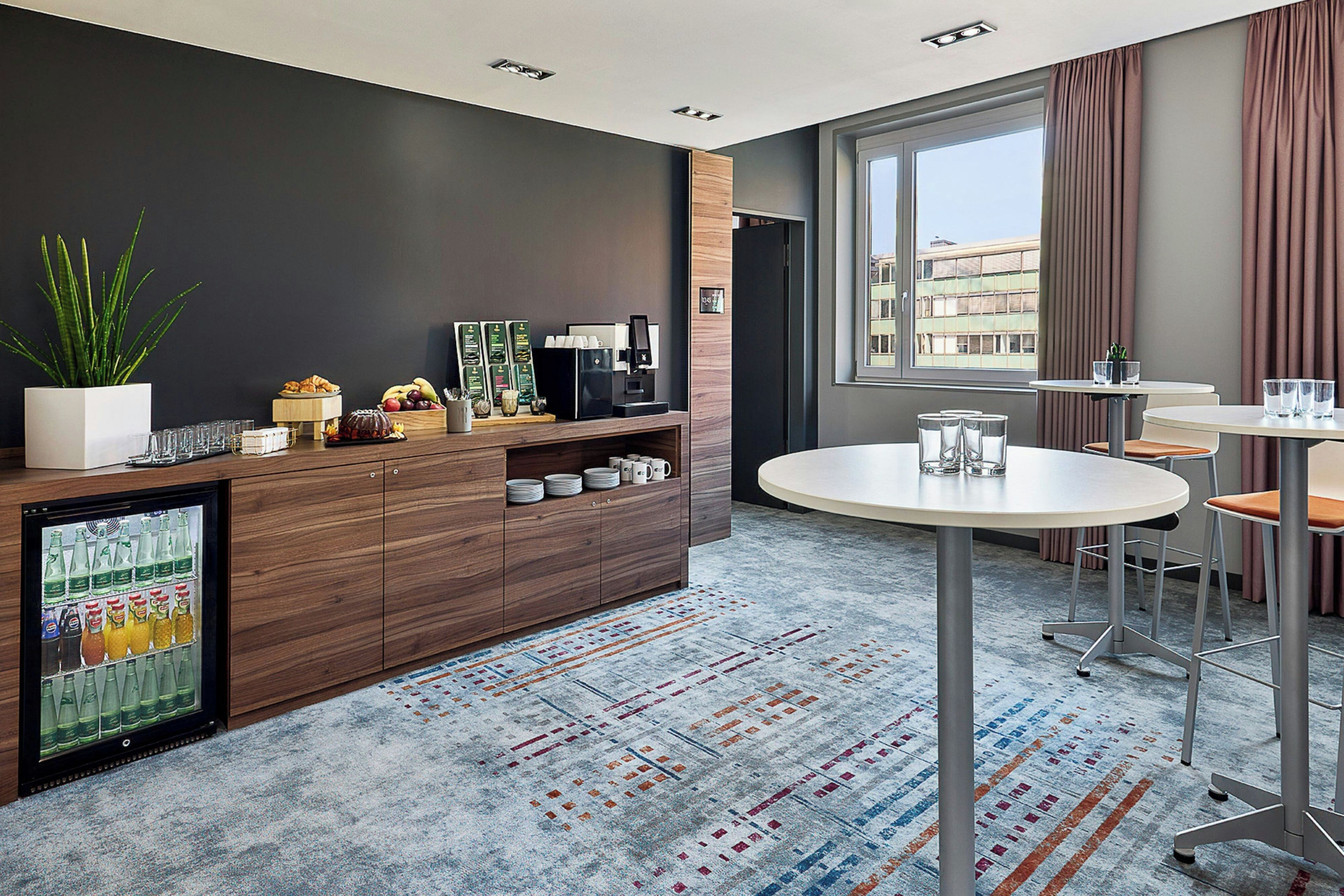 A modern hotel room with a wooden reception desk, a coffee machine, and a refrigerator. There are two round tables with chairs, a large window with curtains, and a patterned carpet.