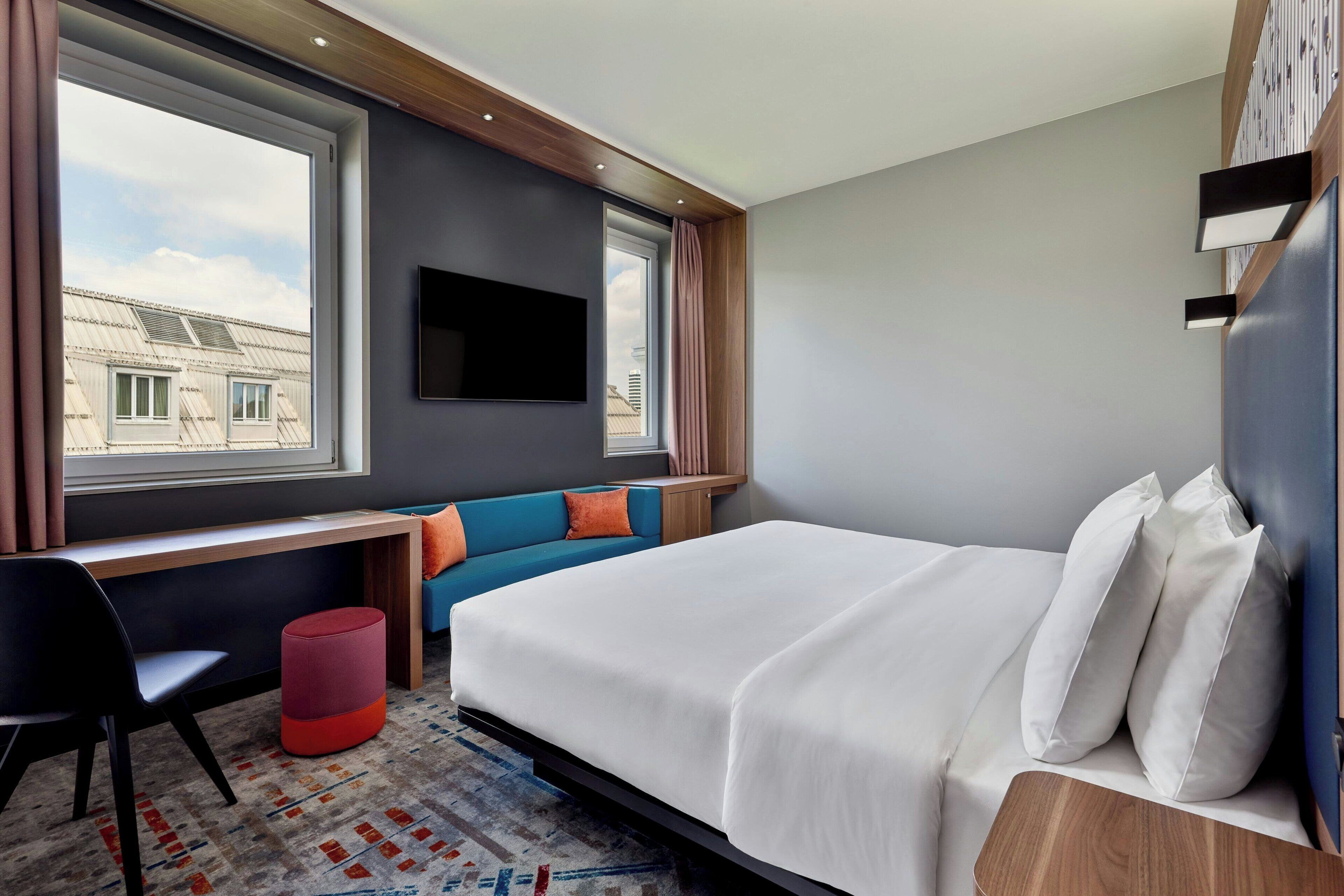 A modern hotel room with a bed, a desk, a chair, a TV, and a window with curtains.