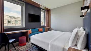 A modern hotel room with a bed, a desk, a chair, a TV, and a window with curtains.