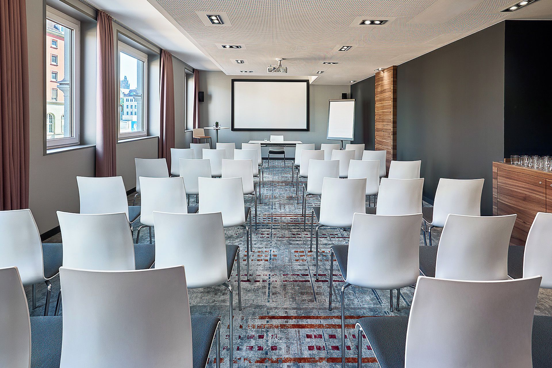 Een vergaderzaal met rijen witte stoelen, een scherm voor een beamer en een groot raam met gordijnen.