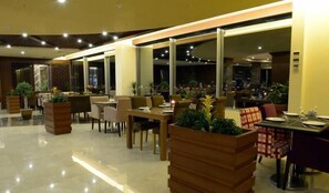 A modern restaurant with indoor seating, large windows, and potted plants. - Mariana Hotel Erbil (Erbil)