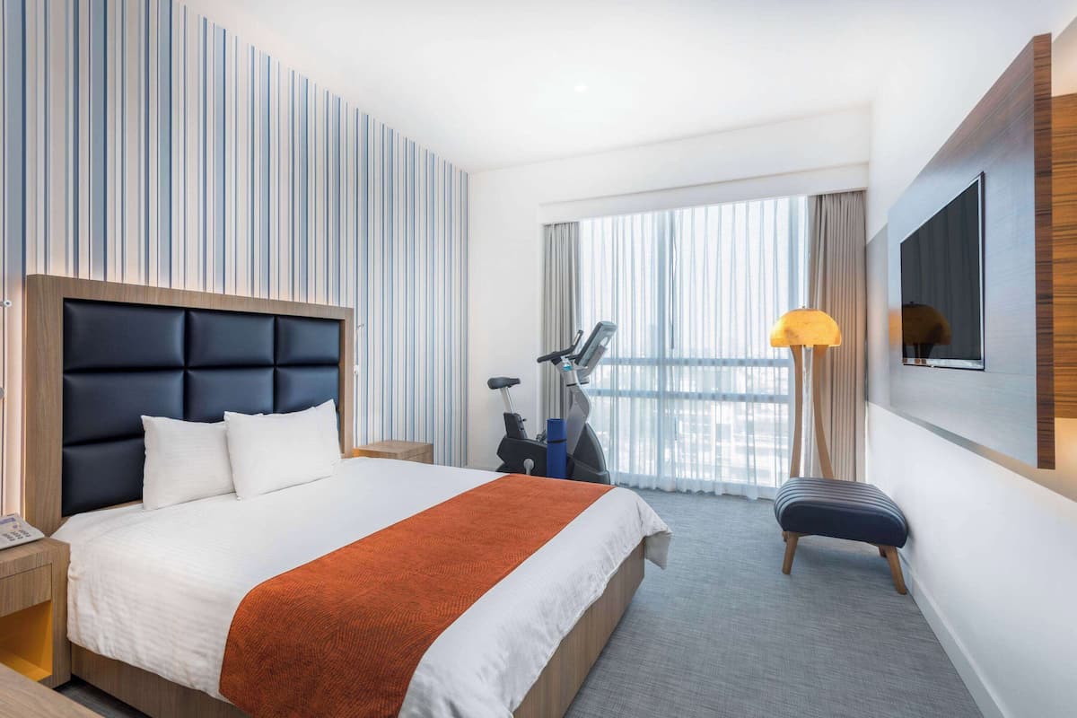 Ein Hotelzimmer mit einem großen Bett, einem Schreibtisch, einem Stuhl, einem Laufrad, einer Stehlampe und einem großen Fenster mit Vorhängen.