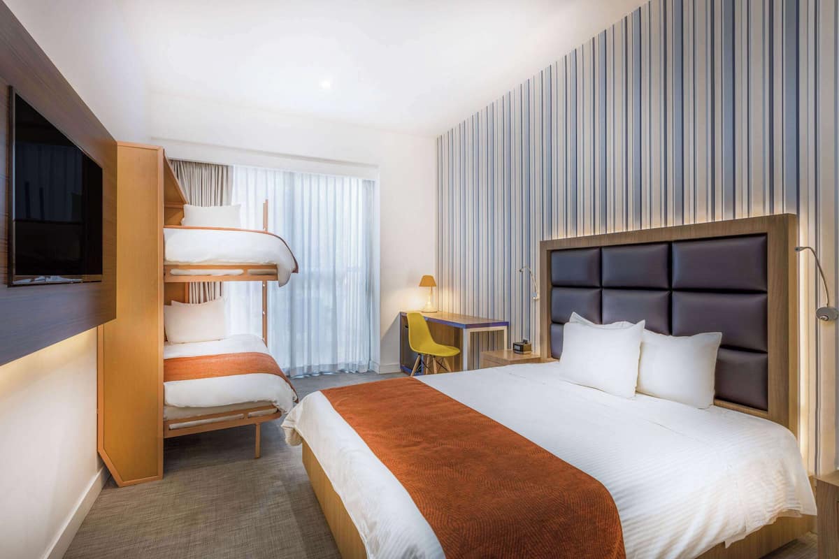 Ein Hotelzimmer mit Etagenbett, Schreibtisch mit Stuhl, Fernseher und einem großen Bett mit Kopfteil.