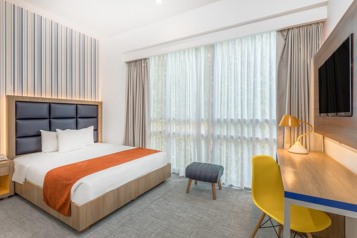 Ein Hotelzimmer mit einem großen Bett, einem Schreibtisch mit Stuhl, einem Fernseher und einem Fenster mit Vorhängen.