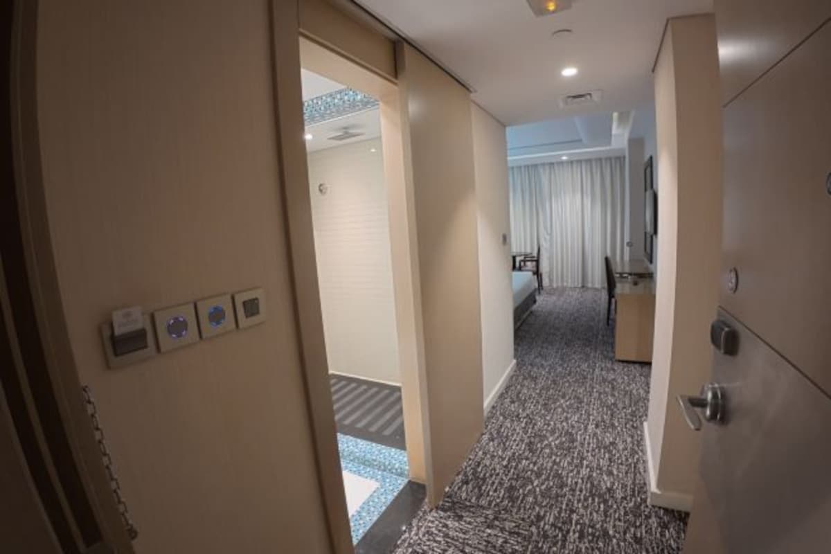A hotel corridor with a carpeted floor, a door leading to a room, and a bed with a white curtain.