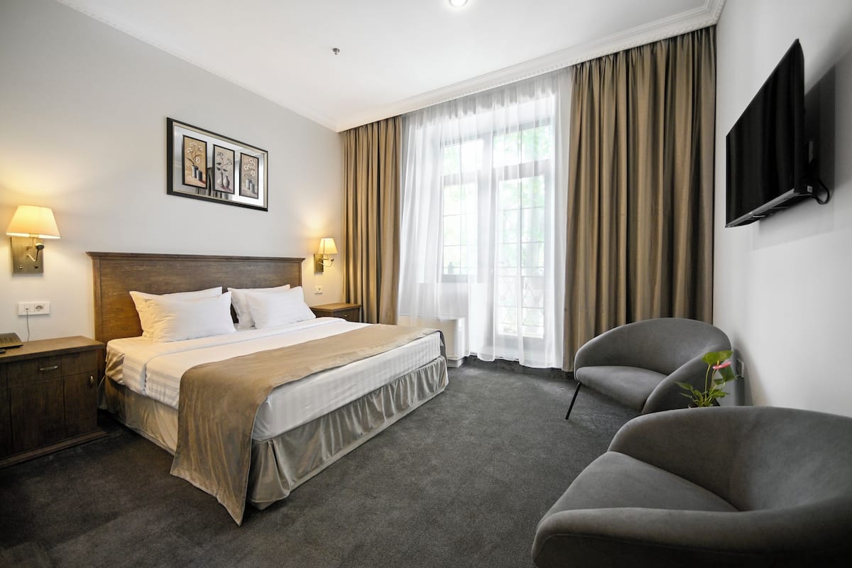 Ein Hotelzimmer mit einem Bett, zwei Nachttischlampen, einem Flachbildfernseher, einem grauen Sessel und einem großen Fenster mit Vorhängen.