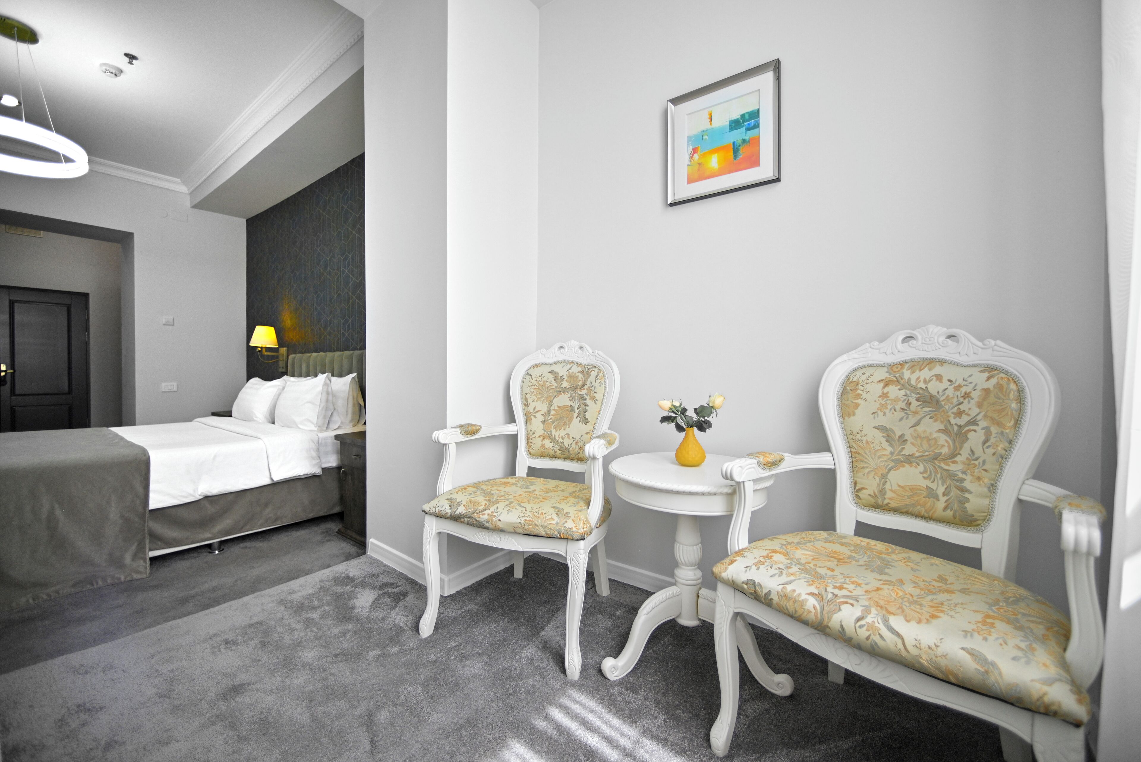 A hotel room with a bed, two chairs, a small table, a vase with flowers, and a framed picture on the wall.