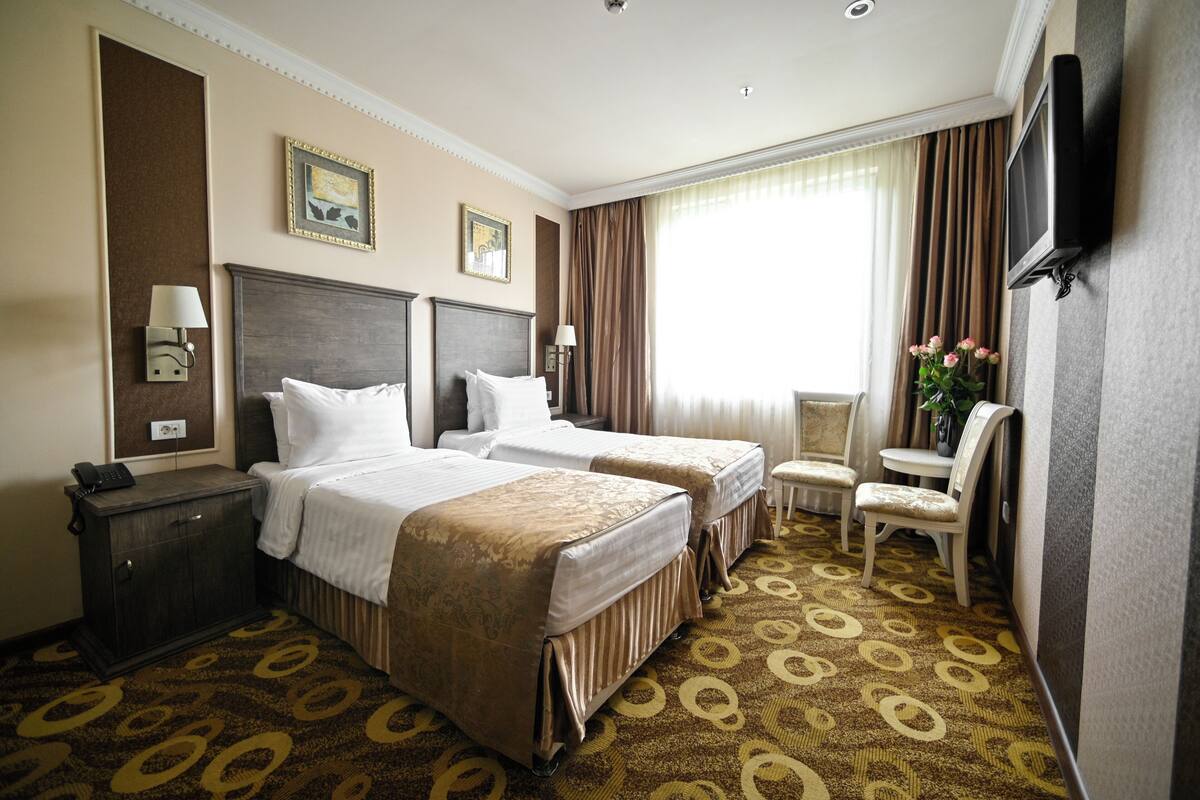 Ein Hotelzimmer mit zwei Betten, einem Schreibtisch mit Lampe, einem an der Wand montierten Fernseher und einem Sessel.