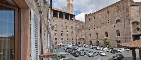 A courtyard with a large building featuring arched windows and a tower. There is a parking lot with several cars.