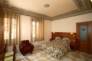 Ein Schlafzimmer mit Schachbrettmuster-Boden, einem Bett mit floralem Bettbezug, einem Ledersessel und einer Holztür mit Glasfenster.
