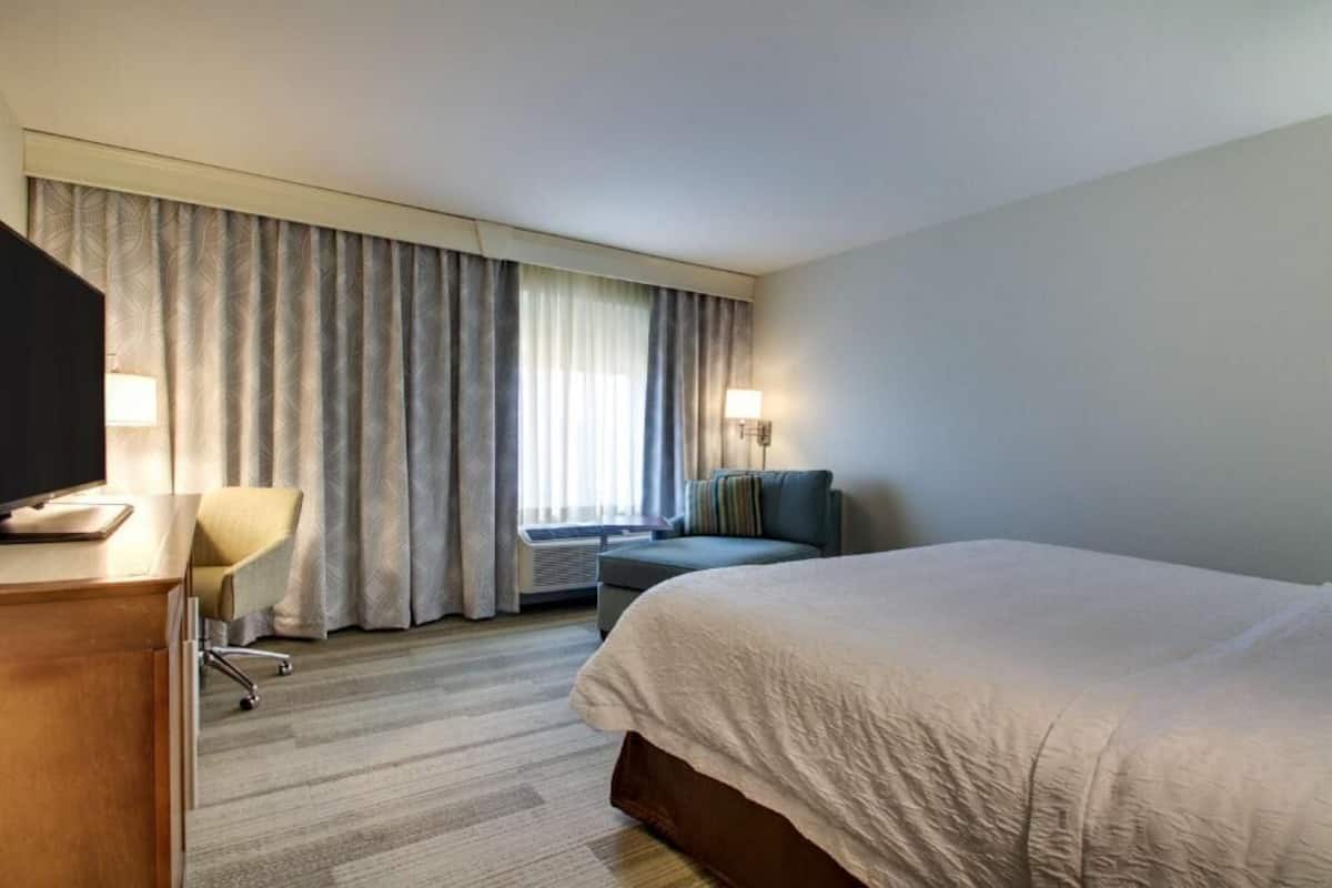 A hotel room with a bed, a desk with a computer, a chair, a sofa, and a window with curtains.