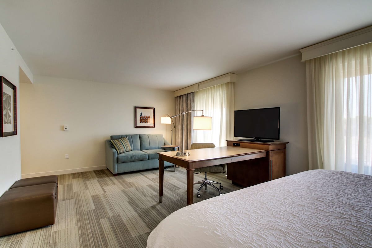 A hotel room with a large bed, a desk with a chair, a sofa, a TV, and a lamp.