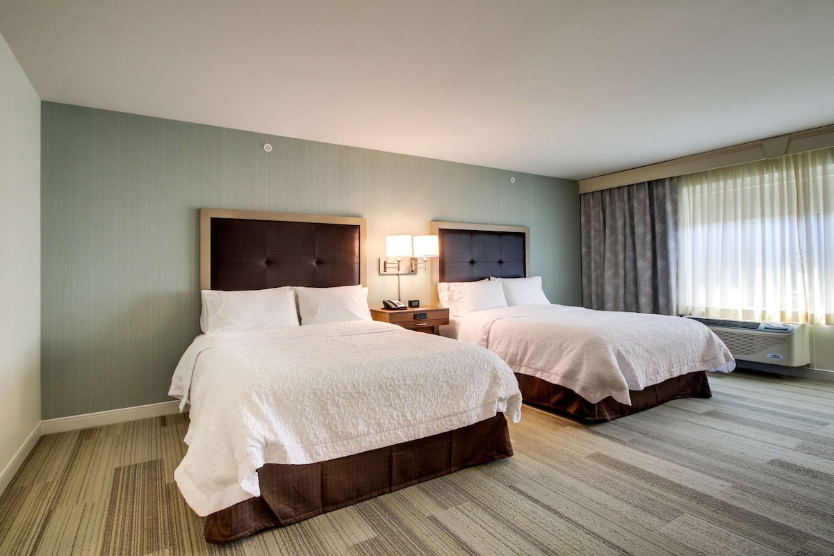 A hotel room with two beds, a nightstand with a lamp, and a large window with curtains.