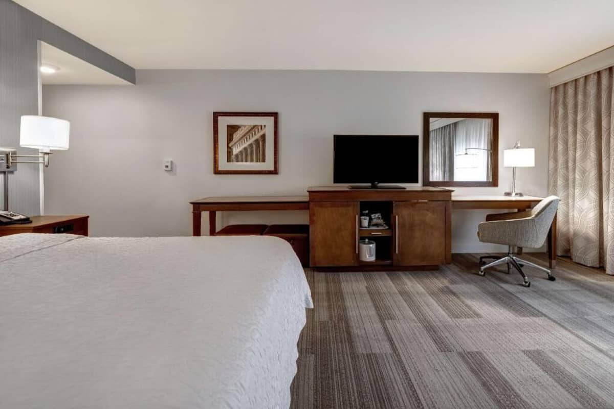 A hotel room with a bed, a desk with a computer, a chair, a television, and a framed picture on the wall.