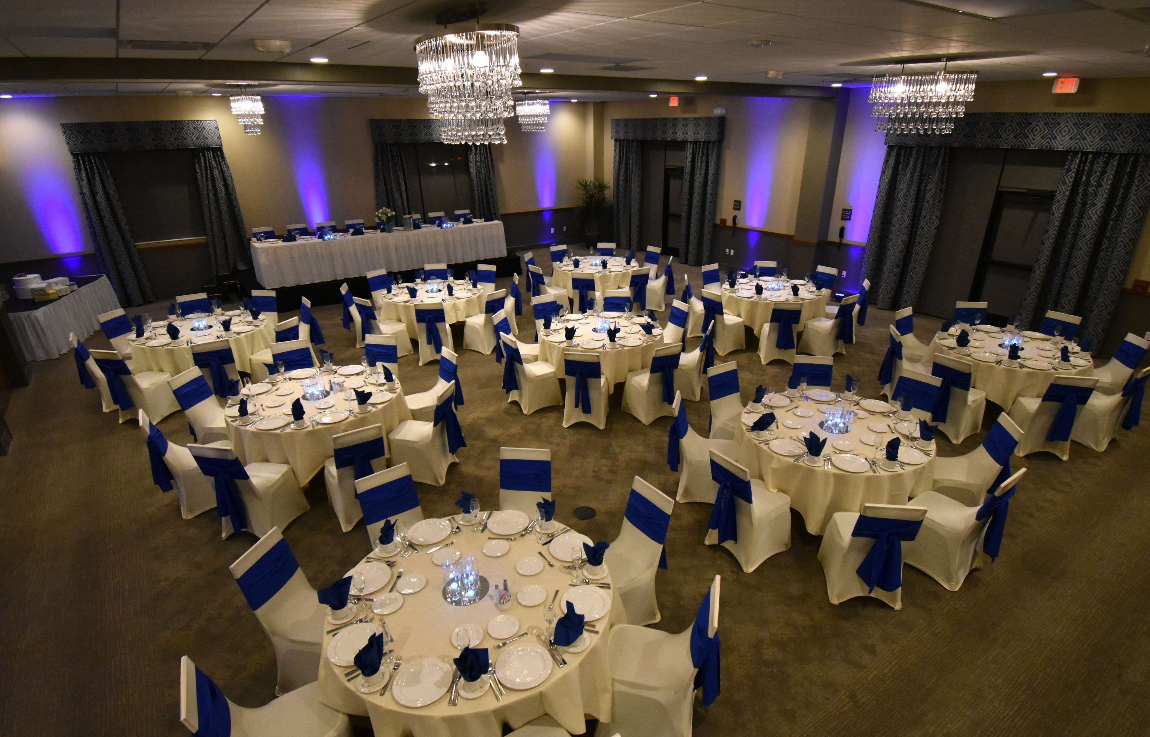 Un salón de banquetes con mesas redondas dispuestas para cenar, cada una con manteles blancos y fajas azules.
