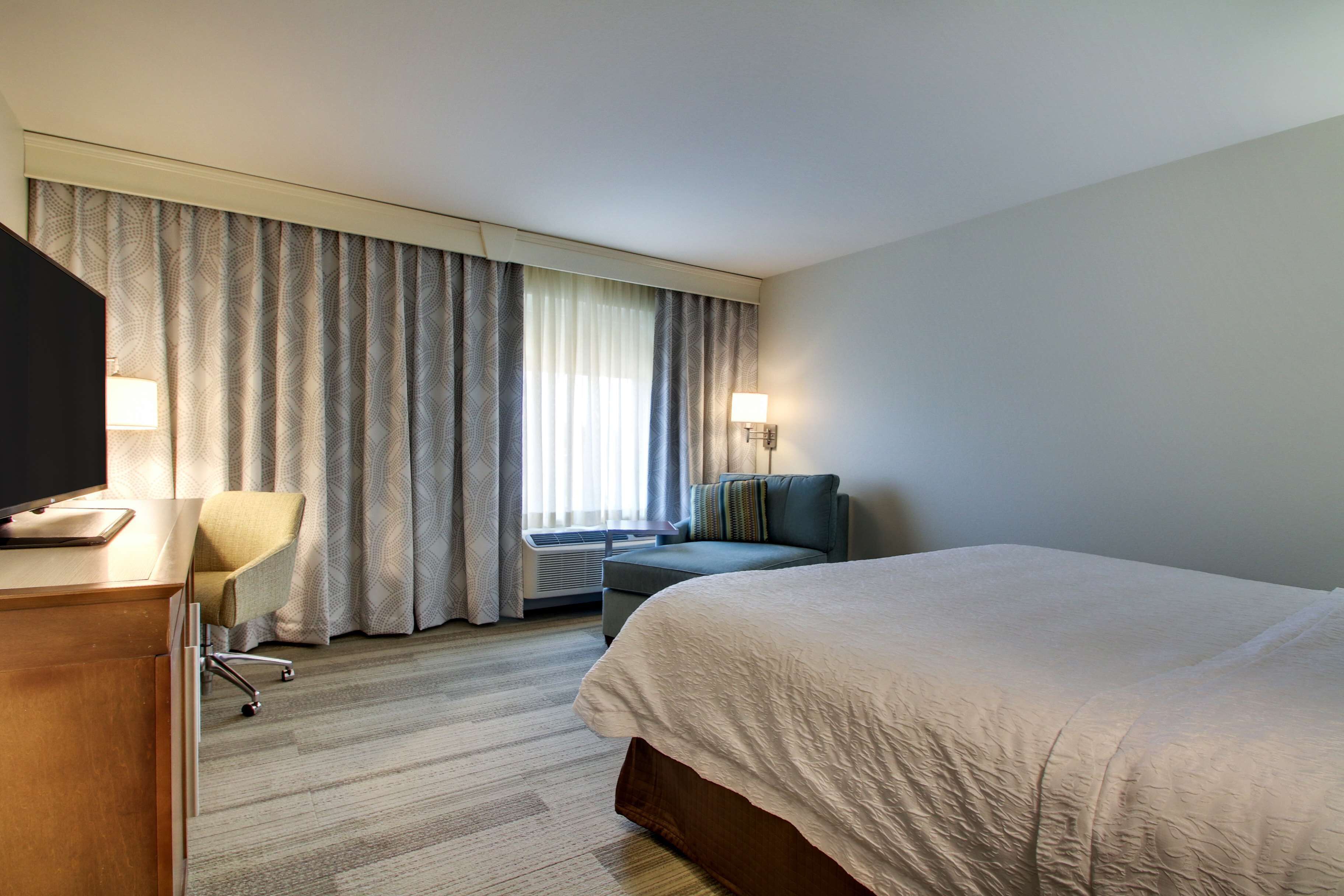 A hotel room with a large bed, a desk with a computer, a chair, a sofa, and a window with curtains.