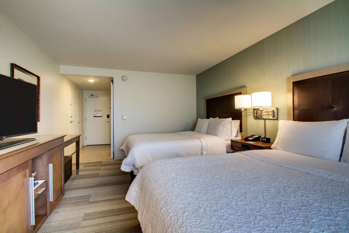 A hotel room with two beds, a desk with a television, a bedside table with a lamp, and a door leading to a bathroom.