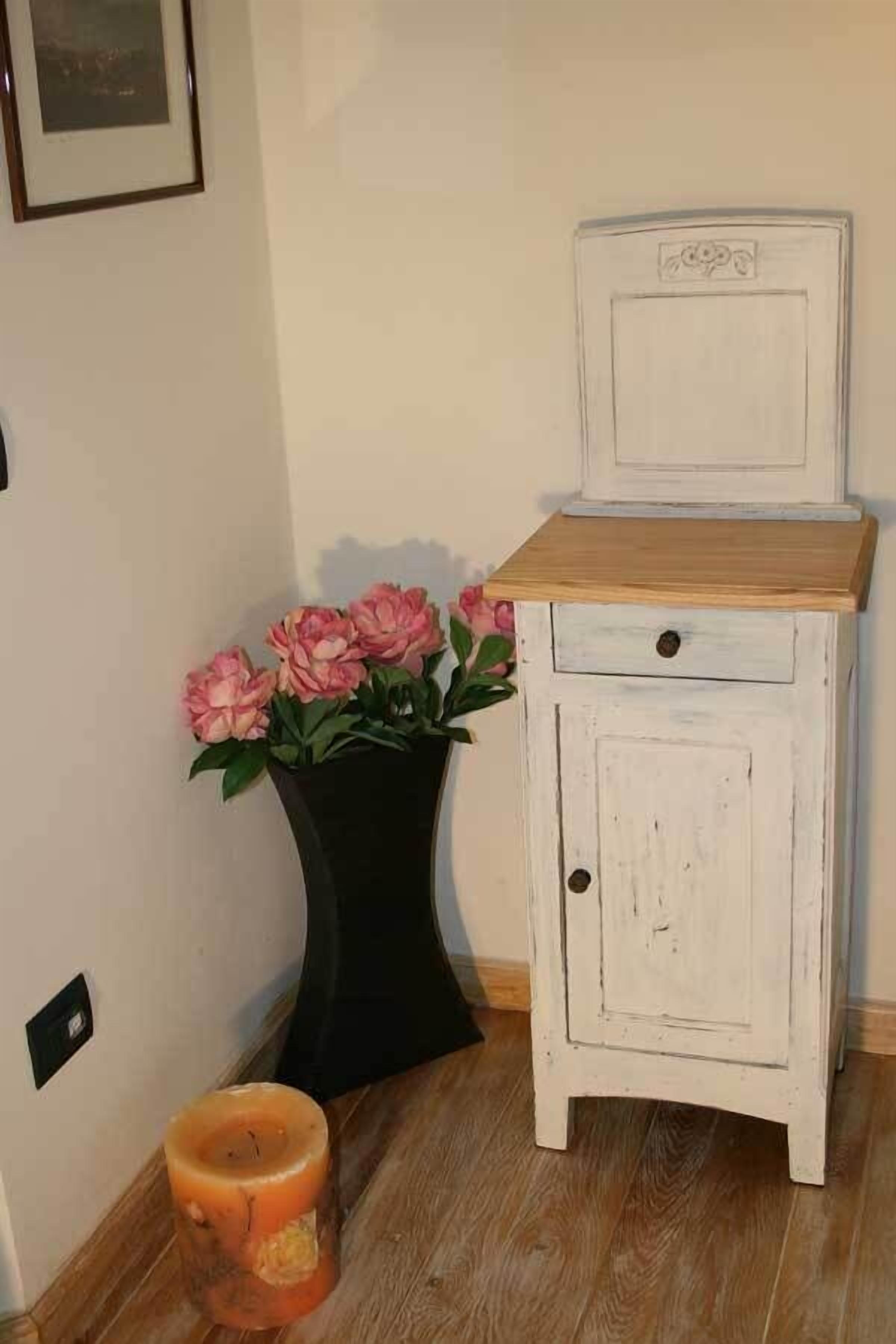 A small white wooden cabinet with a drawer and a shelf, placed next to a black vase with pink flowers on a wooden floor.