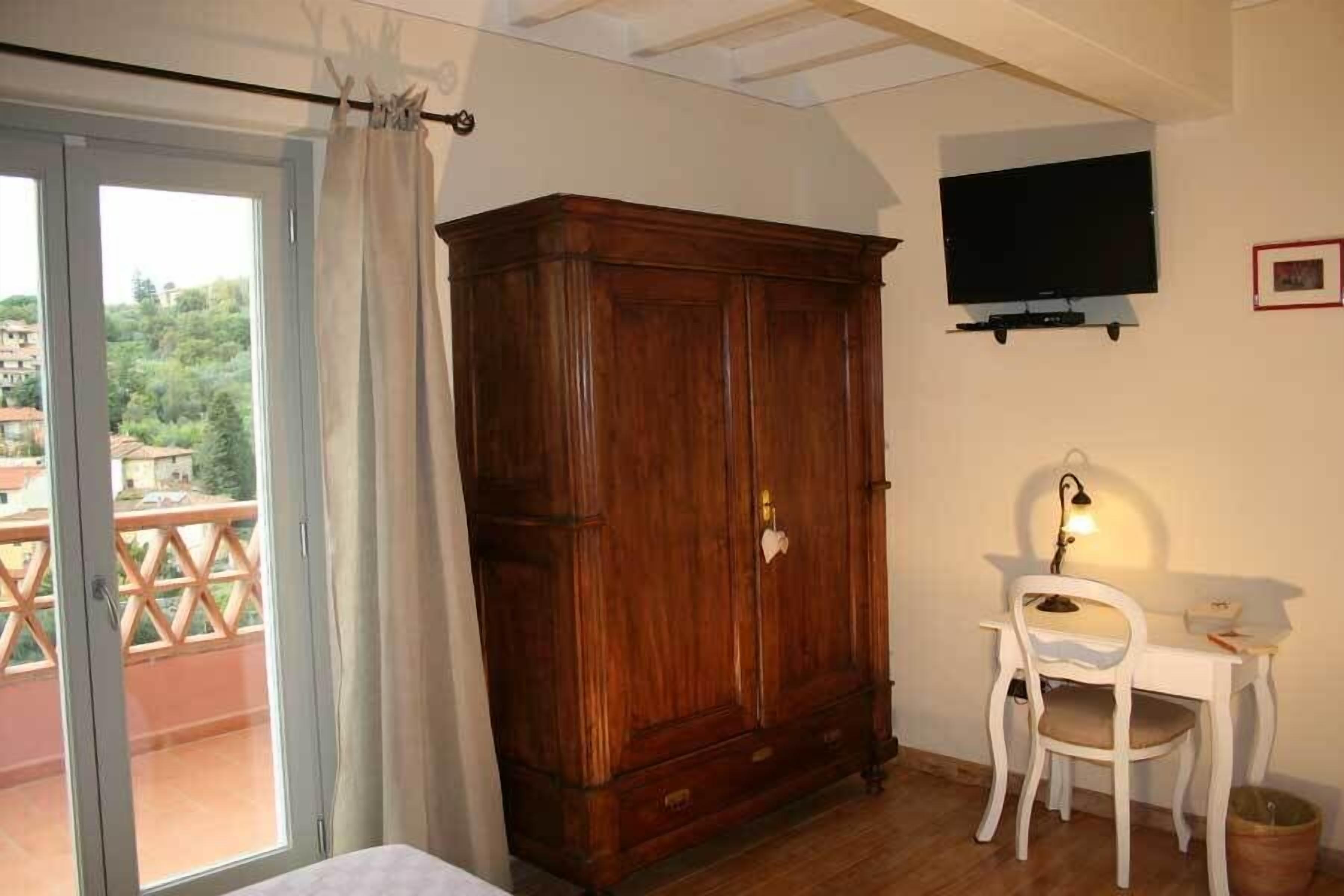 Una habitación con un armario de madera, un escritorio blanco con silla, televisor montado en la pared y un balcón con vista a la vegetación.