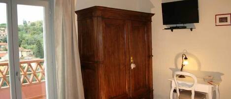 Una habitación con un armario de madera, un escritorio blanco con silla, televisor montado en la pared y un balcón con vista a la vegetación.