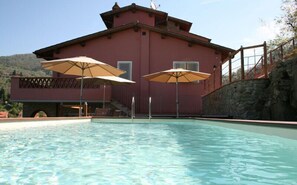 A swimming pool with two umbrellas, a red building, and a stone wall. - La Terrazza di Reggello (Reggello)