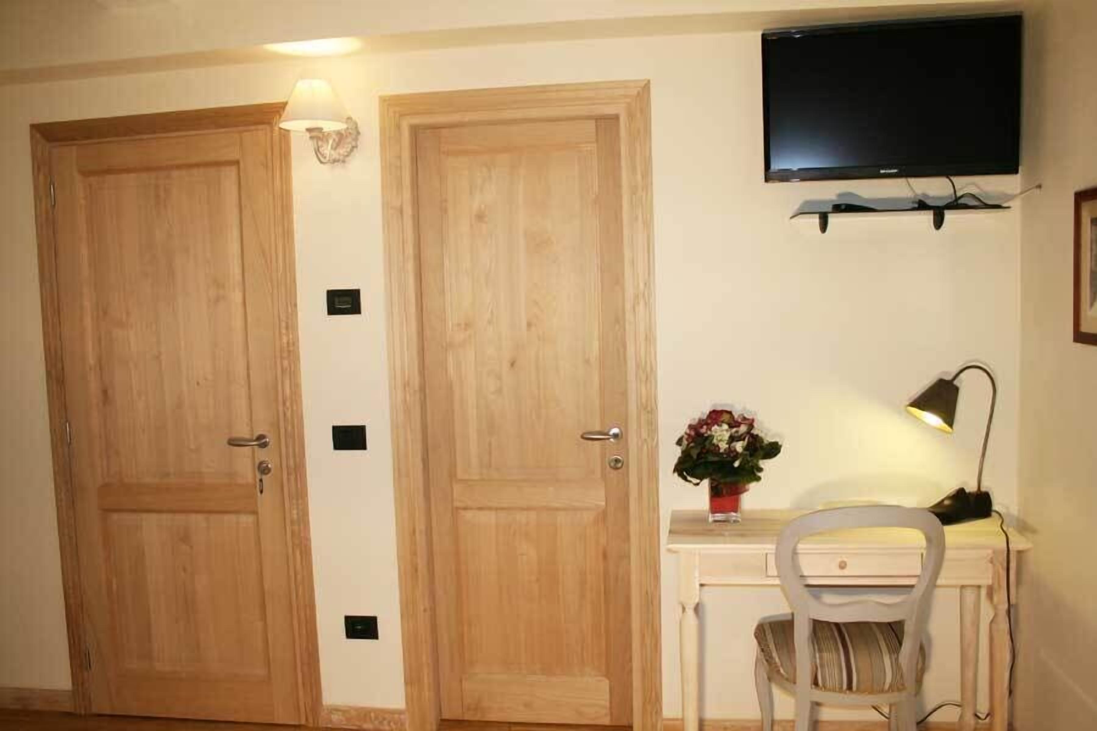 Une pièce avec deux portes en bois, un bureau avec une chaise, un téléviseur fixé au mur et une petite table avec une lampe et un vase à fleurs.