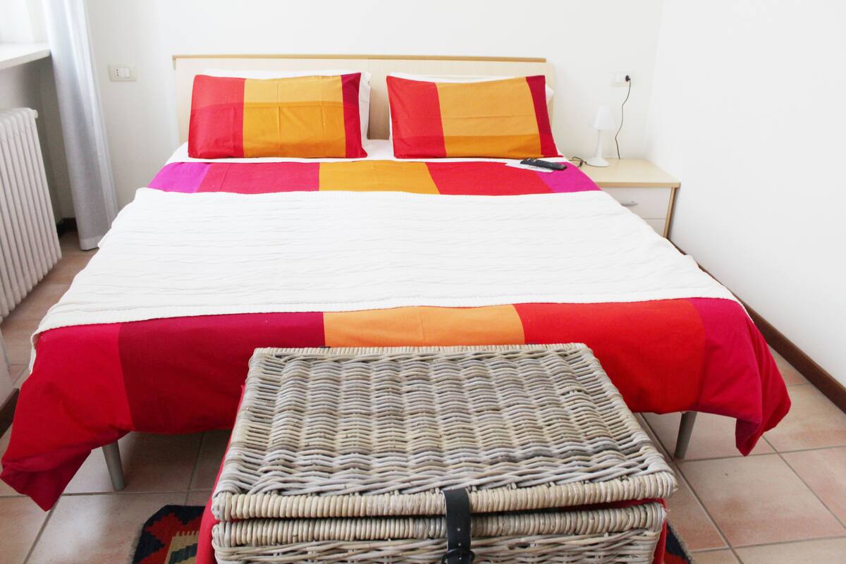 A bed with a red and orange bedspread, a bedside table with a lamp, and a wicker bench.