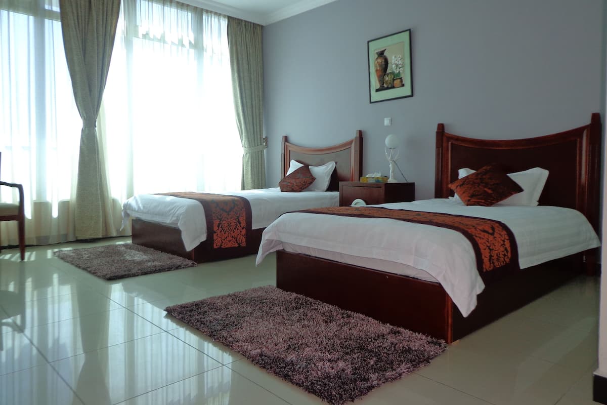 A hotel room with two beds, a nightstand, a lamp, a painting on the wall, and a view of the outdoors through the window.