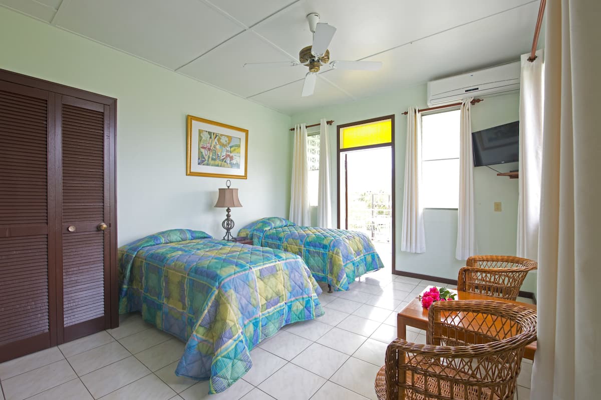 Quarto de hotel com duas camas, ventoinha no teto, ar condicionado, televisão, mobiliário de vime e um quadro na parede.