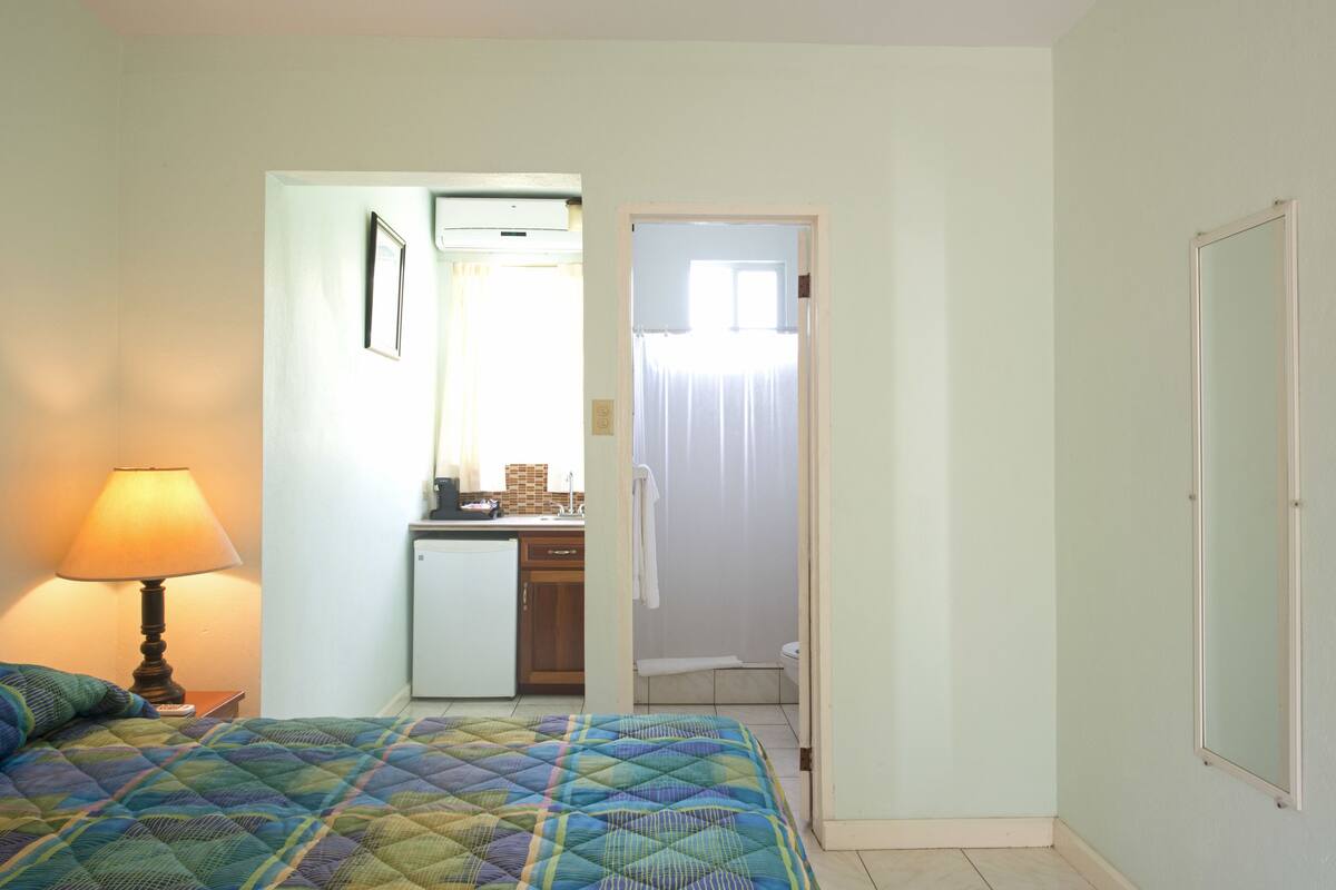 Quarto com cama, luminária de cabeceira, uma kitchenette pequena e banheiro visível por uma porta aberta.