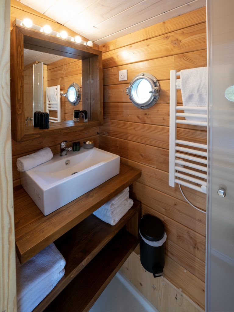Une salle de bain rustique, avec des murs en bois, un lavabo blanc, deux miroirs, un porte-serviettes et une poubelle.