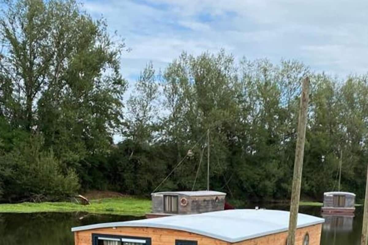 Een houten woonboot met een plat dak, een klein dek en een raam met gordijnen.