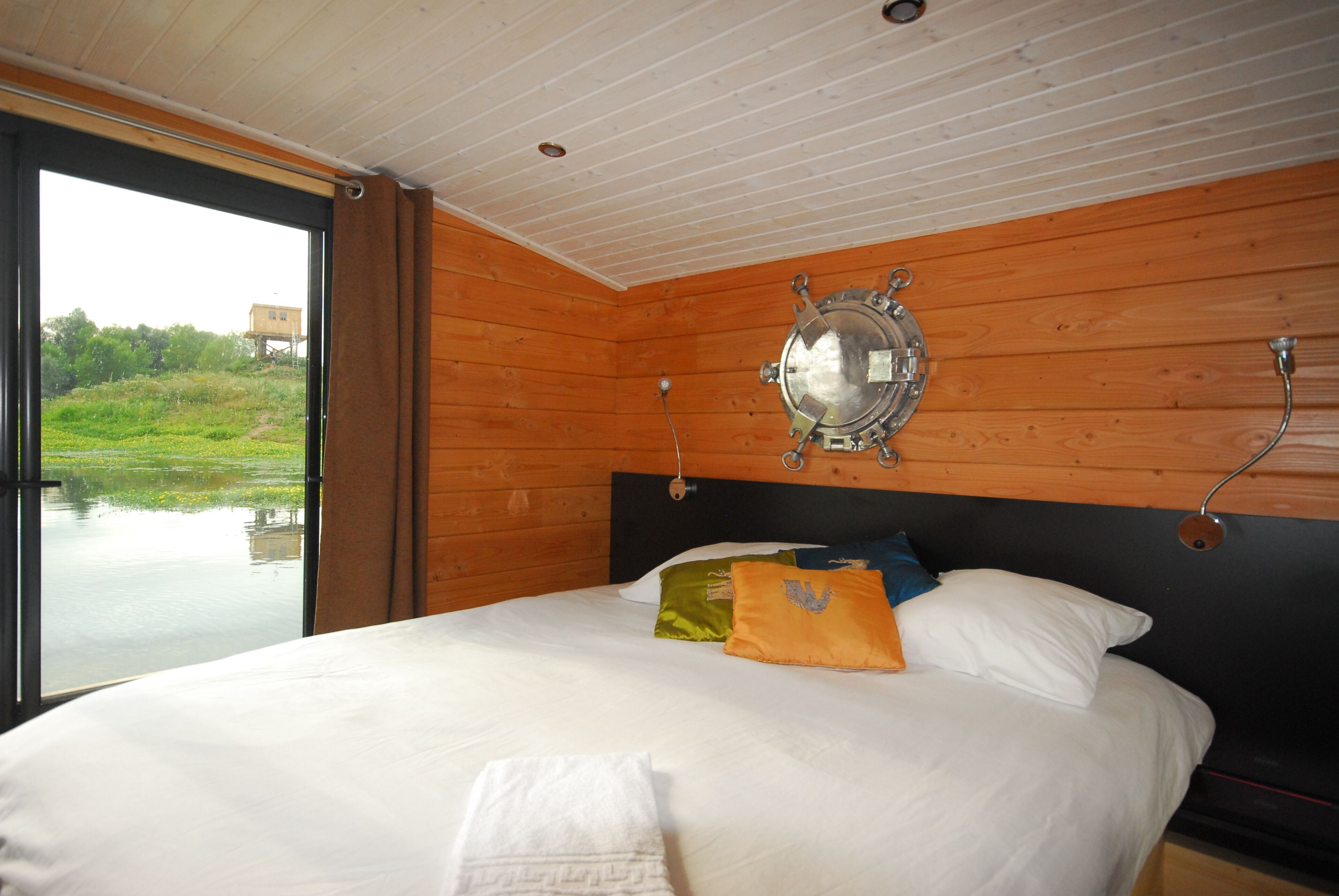 A bedroom with a large bed, wooden walls, a view of a water body, and a ship's wheel decoration.