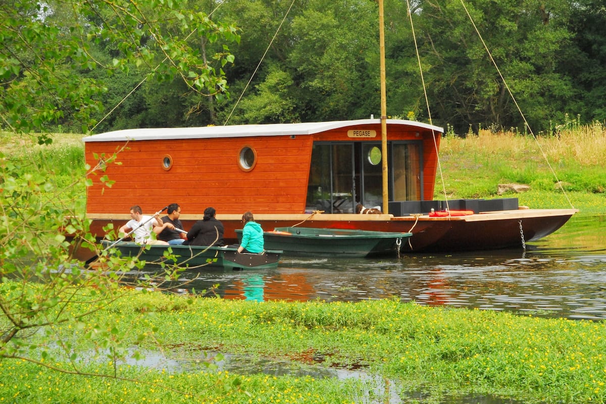 Een houten woonboot met een hutje en een klein dek, omgeven door water en groen.