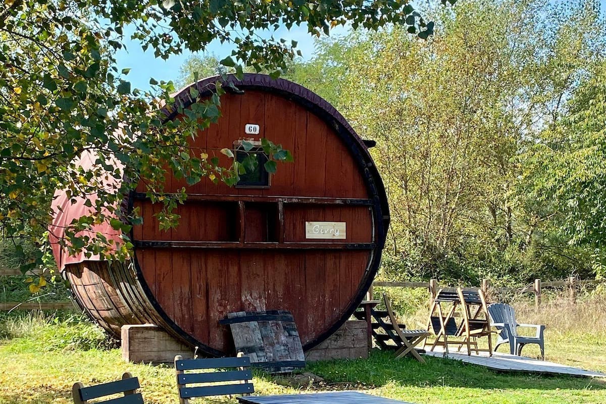 Een rustieke buitenruimte met een houten tafel en stoelen, een grote tonconstructie en een houten terras.