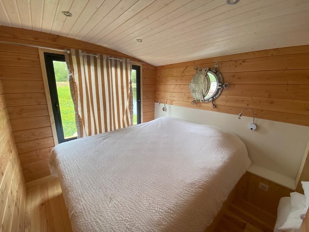 A wooden cabin bedroom with a large bed, a window with blinds, and a round mirror on the wall.