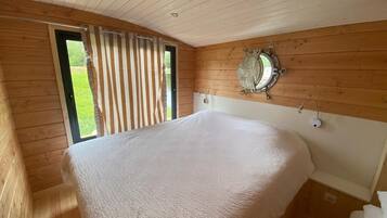 A wooden cabin bedroom with a large bed, a window with blinds, and a round mirror on the wall.