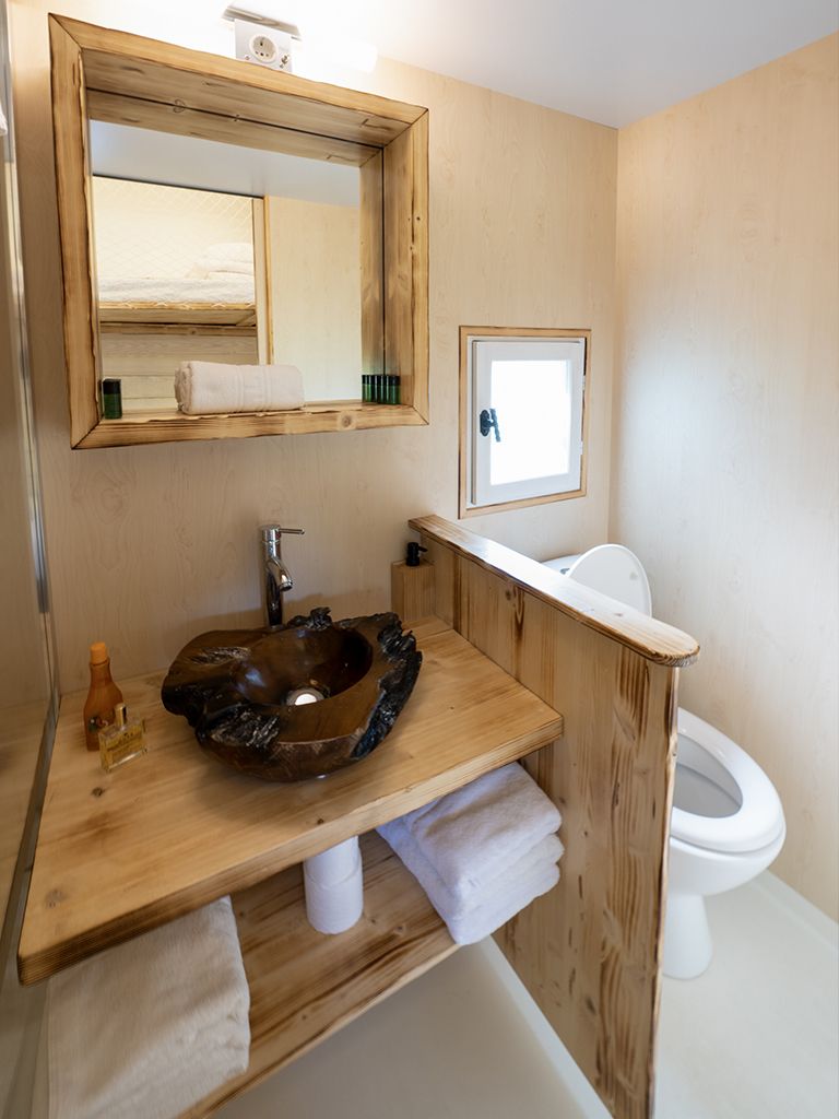 Un baño con tocador de madera, un espejo grande, lavamanos con un lavabo original, inodoro y una ventana pequeña.