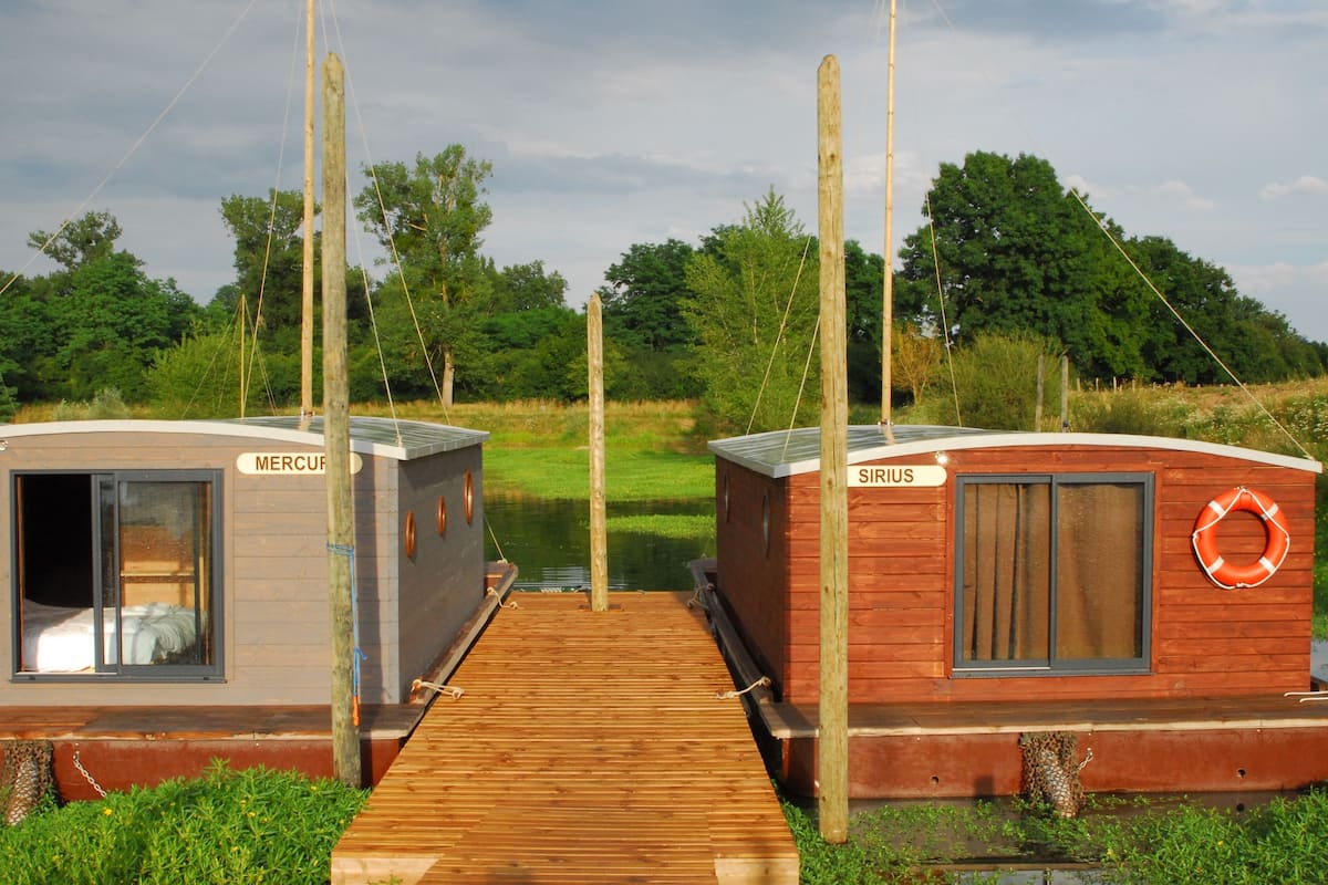 Twee houten hutten, genaamd 'Mercury' en 'Sirius', met een houten steiger daartussen, gelegen voor een achtergrond van groen en bomen.