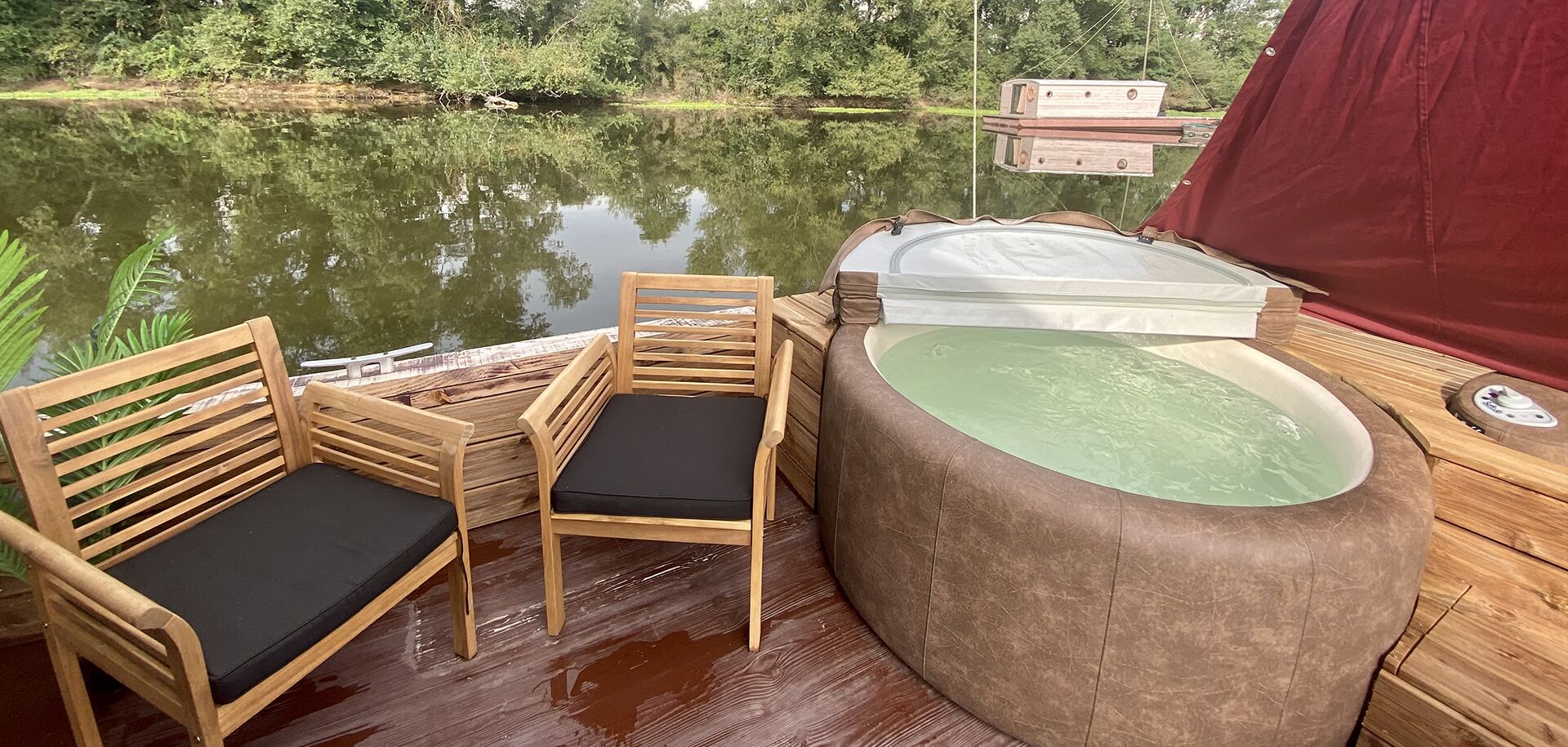 A wooden deck with two wooden chairs with black cushions, a round wooden tub with a green interior, and a red canopy.