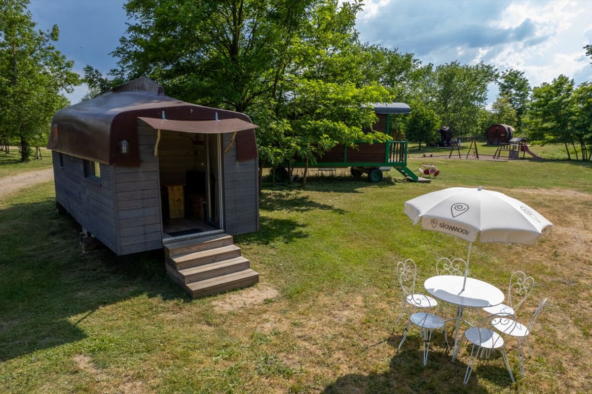 Een klein houten vakantiehuisje met een overdekte veranda, een picknicktafel met stoelen, en een speeltuin op de achtergrond.