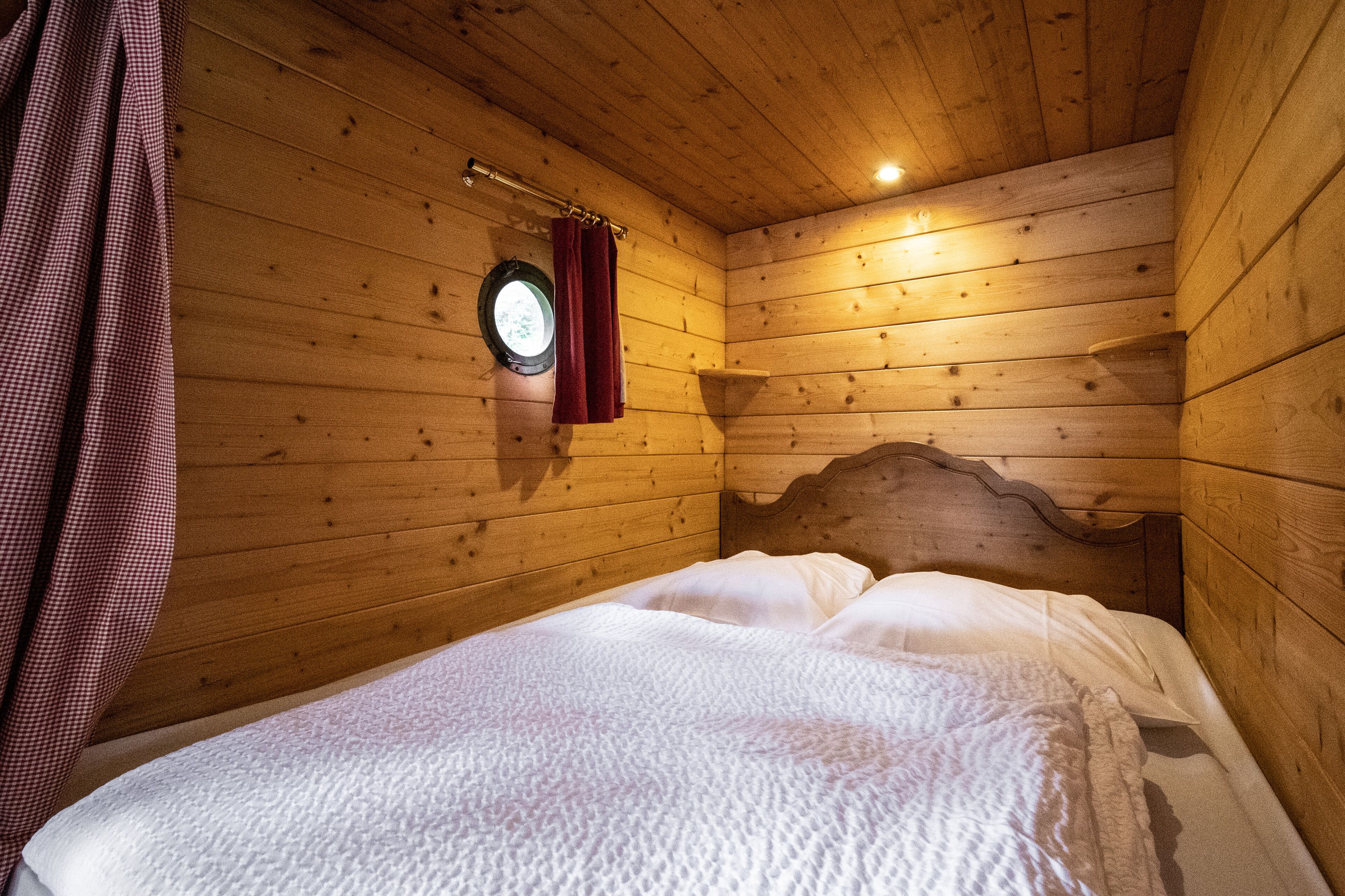 A wooden cabin room with a bed, wooden walls, a window with curtains, and a headboard.