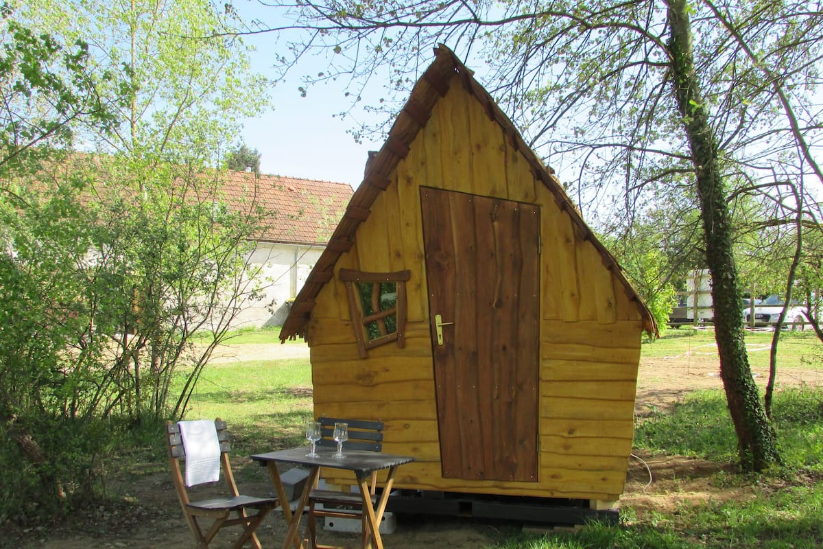 Een klein houten vakantiehuisje met een deur en raam, een tafel met twee stoelen en een bank.
