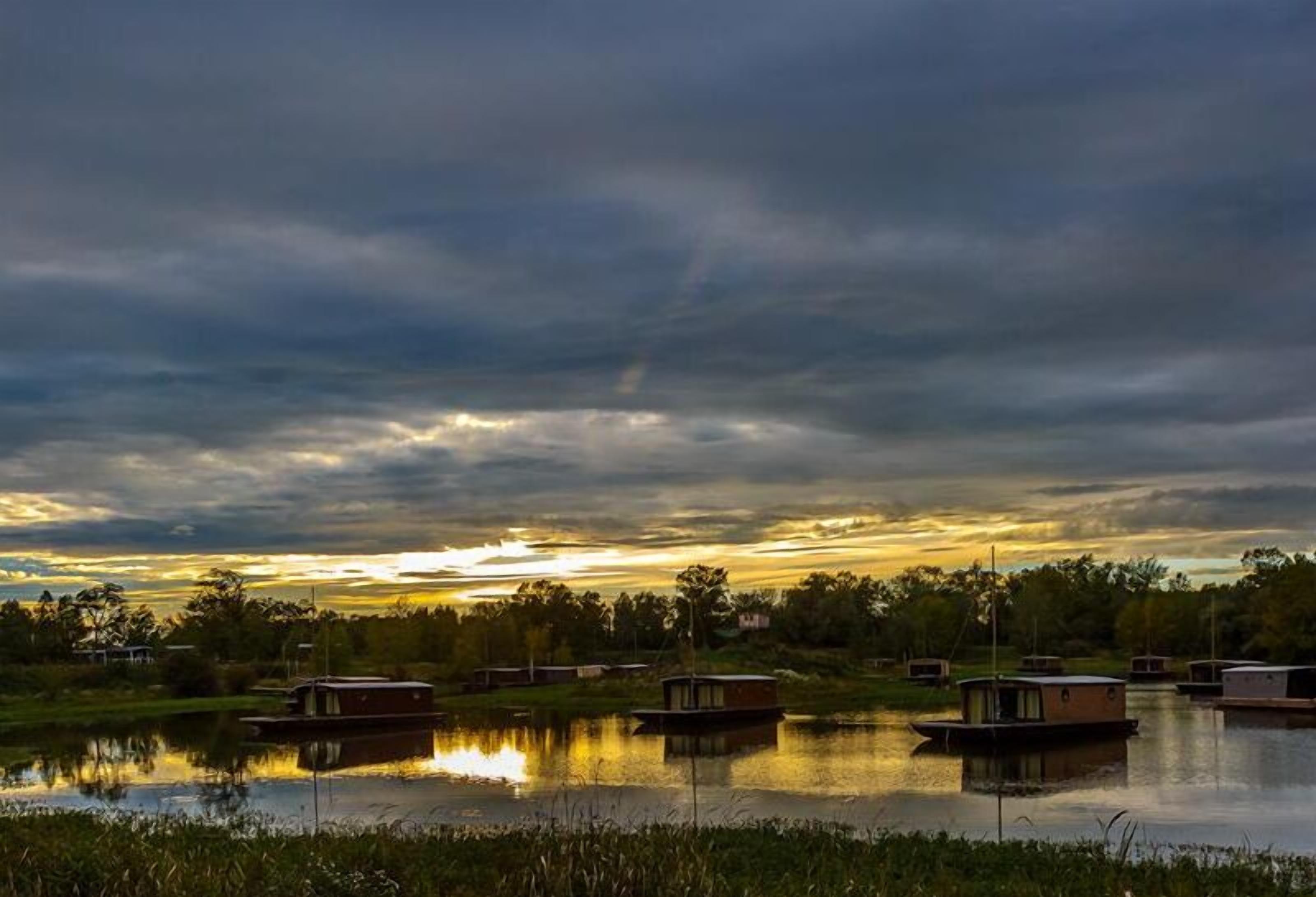 A calm lake with several floating structures, likely used for accommodation, surrounded by greenery and under a cloudy sky.