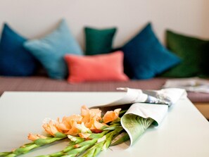 Un ramo de flores sobre una mesa blanca con una revista, frente a un sofá con almohadones de colores.