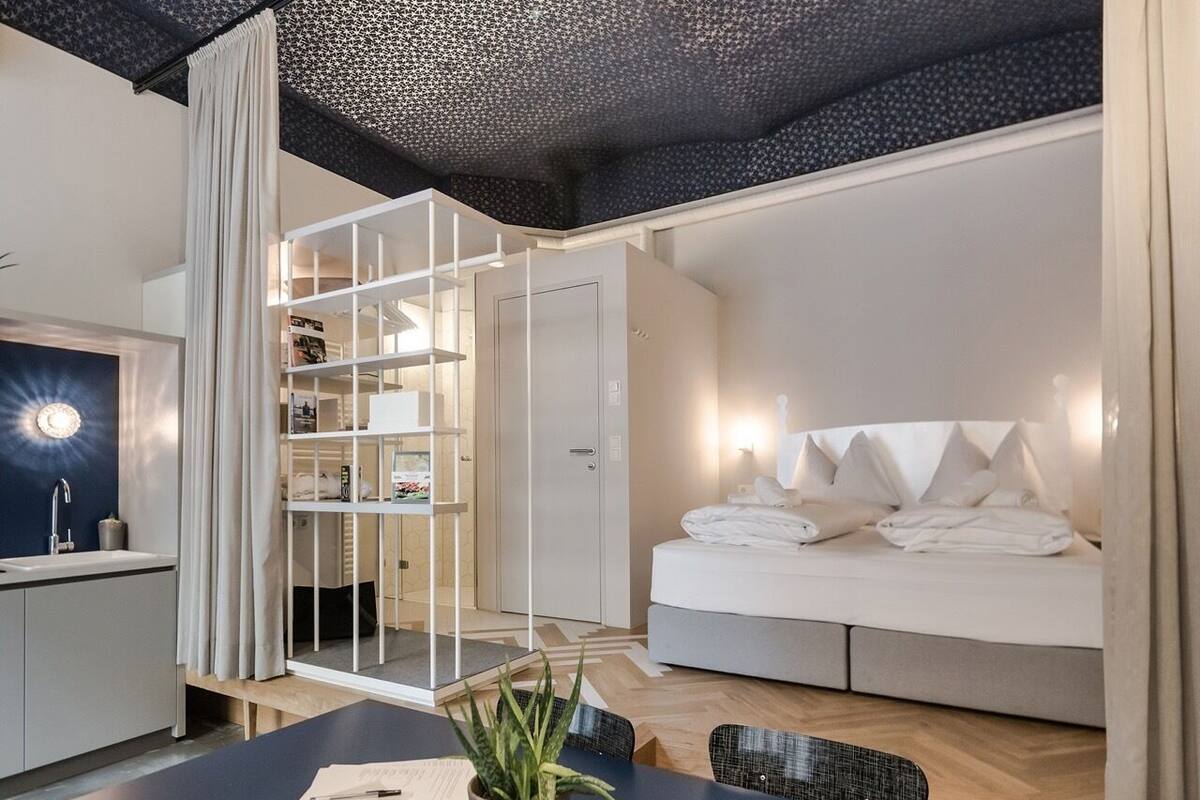 A modern hotel room with a bed, a desk, a chair, a plant, and a wardrobe.